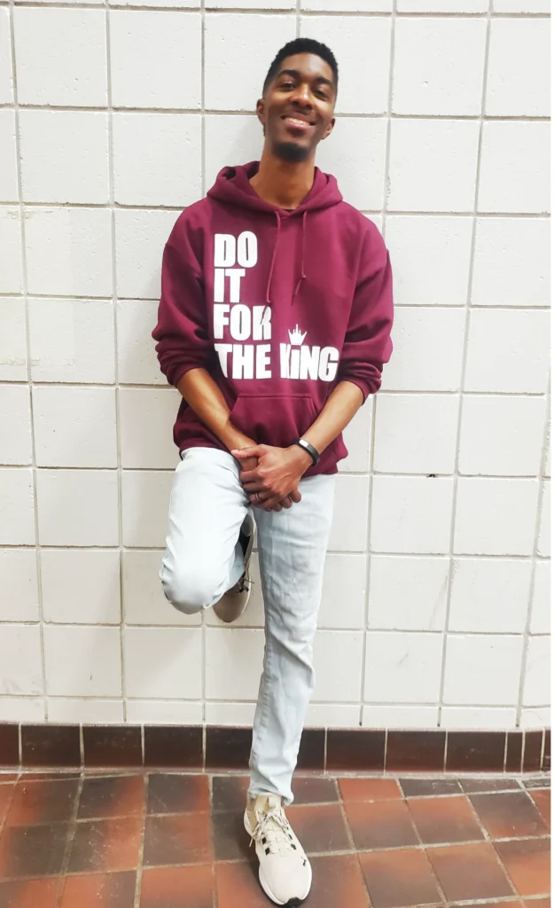 Customer wearing a maroon “Do It For The King” hoodie, standing against a tiled wall with hands clasped and smiling.